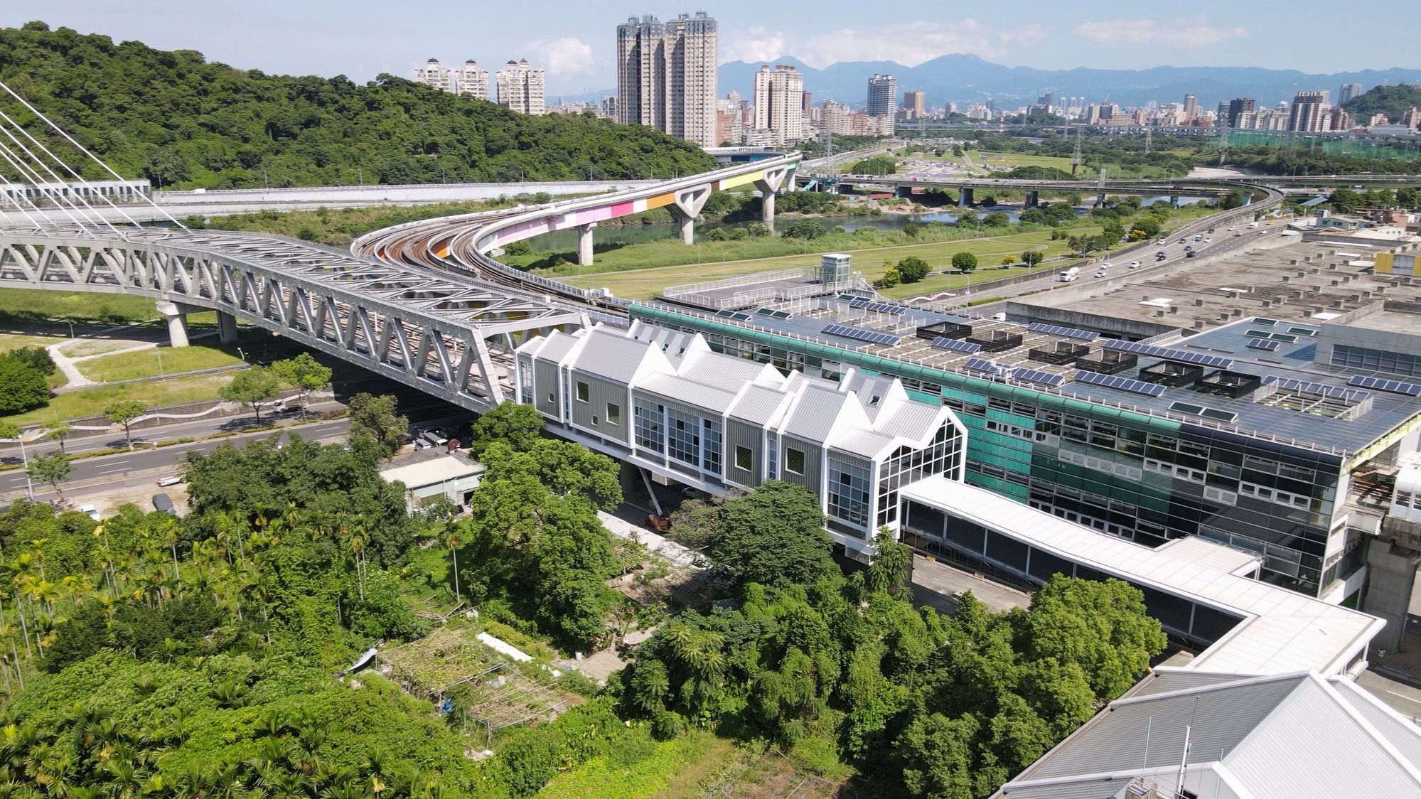 從空中看光合創學所在的建築:一排白色山形屋頂,左側是環狀線十四張站的高架橋與軌道,遠方是台北市區與群山